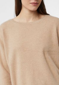 Kobieta ubrana w miękki beżowy sweter z okrągłym dekoltem, widoczna od ramion do górnej części klatki piersiowej, z długimi brązowymi włosami.