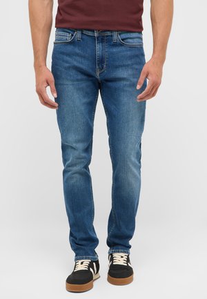Mann trägt blaue Slim-Fit-Jeans und schwarze Sneakers mit weißen Schnürsenkeln und steht vor einem einfarbigen hellen Hintergrund.