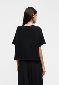 Femme aux longs cheveux noirs portant un haut ample noir à manches courtes et un pantalon noir, vue de dos sur un fond blanc uni.