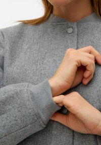 Chaqueta gris de textura suave con puños acanalados y cuello redondeado. Presenta un cierre de botón simple cerca del escote.