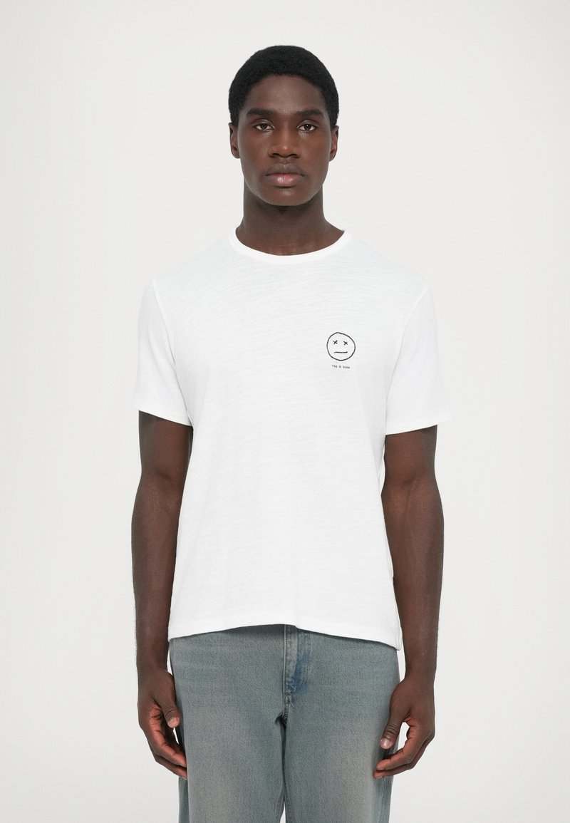 White cotton t-shirt with short sleeves, round neck, featuring a small black smiley face graphic on the chest. Pair of light blue jeans.