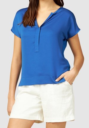 Blouse bleue à manches courtes en tissu lisse, dotée d'un col en V avec patte de boutonnage, associée à un short blanc. Détails texturés sur l'ourlet.