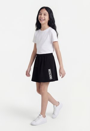 Fille souriante marchant, portant un t-shirt blanc, une jupe noire avec le texte "MOSCHINO" et des baskets blanches sur un fond blanc uni.