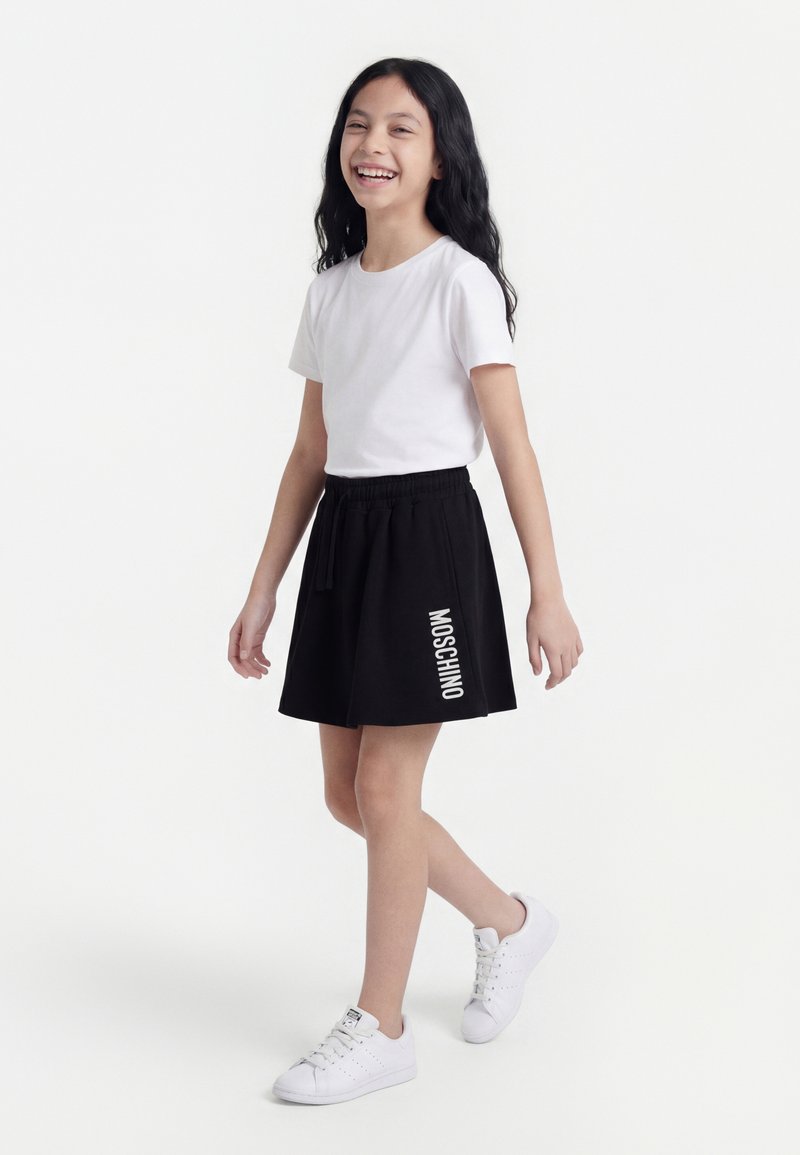 Fille souriante marchant, portant un t-shirt blanc, une jupe noire avec le texte "MOSCHINO" et des baskets blanches sur un fond blanc uni.