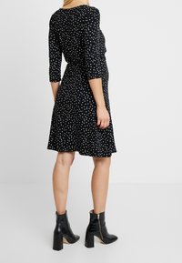 Kobieta ubrana w czarną sukienkę do kolan w białe i niebieskie kropki, w połączeniu z czarnymi skórzanymi botkami na obcasie, stojąca na tle białym.