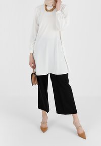 Weißes Langarmoberteil kombiniert mit schwarzen, weit geschnittenen Hosen, die ein strukturiertes, fließendes Design aufweisen. Accessoires sind eine kleine braune Handtasche und beigefarbene Absätze.