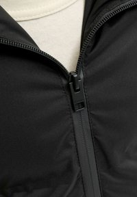 Giacca nera con zip, realizzata in tessuto liscio, con cerniera frontale e tiretto nero. Il rivestimento interno è di colore beige chiaro.