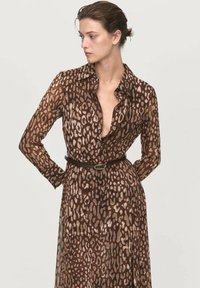 Mujer con un vestido de botones de estampado de leopardo marrón y un cinturón negro, mirando hacia un lado con las manos detrás de la espalda, contra un fondo liso.