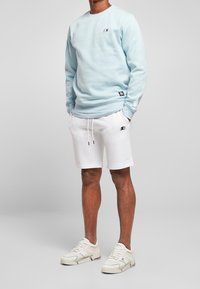 Sudadera azul claro con cuello redondo y logo negro, combinada con pantalones cortos blancos con cordón y logo. Zapatillas blancas completan el conjunto.