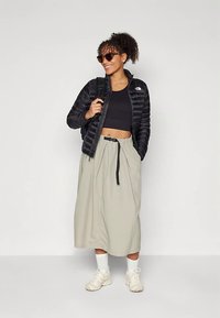 Černá péřová bunda přes černý crop top, doplněná béžovou sukní s vysokým pasem. Bílá ponožky a světlé tenisky dokončují outfit.