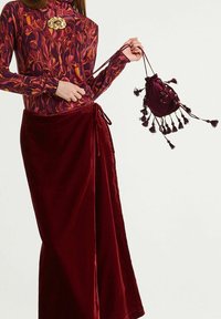 Samtene Wickelrock in Burgunderrot, kombiniert mit einem gemusterten Langarmoberteil. Das Oberteil zeigt florale Designs in Rot-, Orange- und Lilatönen. Eine Kordelzugtasche mit Quasten rundet das Outfit ab.