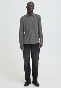 Chemise grise à manches longues avec boutons et une poche poitrine, associée à un jean droit en denim foncé et des chaussures noires.