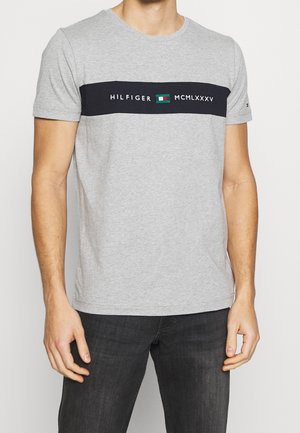 Gray cotton t-shirt featuring a black horizontal band with "HILFIGER" and "MCMLXXXV" text, alongside a colored logo patch. Crew neck design.
