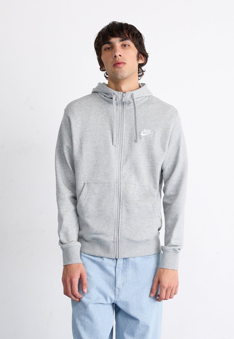 Sudadera gris con cremallera y bolsillo frontal, con cordones. Presenta un pequeño logotipo blanco de Nike en el pecho. La tela parece suave y lisa.