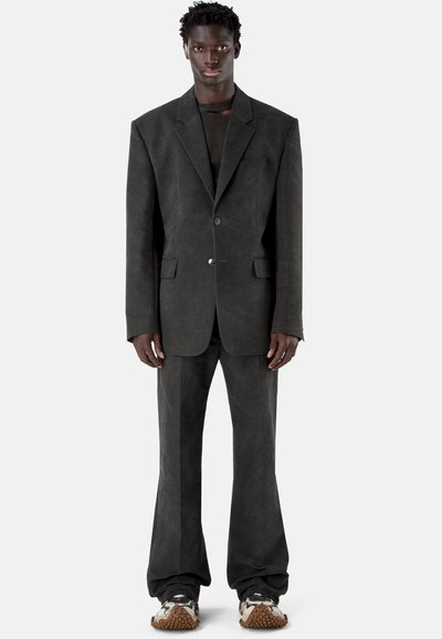 Wysoka osoba stojąca, ubrana w za duży ciemnoszary garnitur z lekko rozszerzanymi spodniami oraz czarno-białe sneakersy z teksturą na podeszwach.