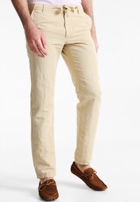 Světlé béžové bavlněné kalhoty s volným střihem, s elastickým pasem na šňůrku a jemnou texturou, v kombinaci s hnědými mokasínovými botami.