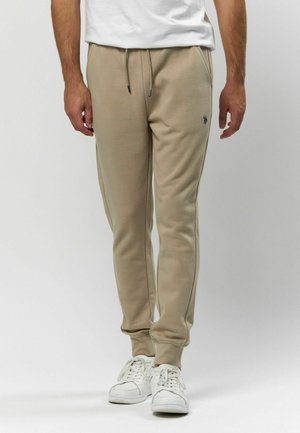 Beige bavlněné joggery s bočními kapsami, žebrovanými manžetami a jemným logem na stehně, doplněné bílými teniskami.