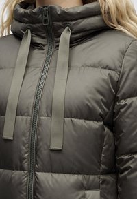 Κοντινό πλάνο σκούρου ελαιόχρωμου επενδυμένου μπουφάν με φερμουάρ και μεγάλες επίπεδες κορδόνια στη κουκούλα, φορεμένο από ένα άτομο με ξανθά μαλλιά.