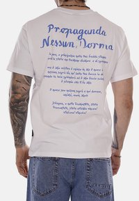 Uomo con maglietta bianca con scritta blu italiana "Propaganda Nessun Dorma" e testo calligrafico, abbinata a jeans blu, che mostra il braccio sinistro tatuato.