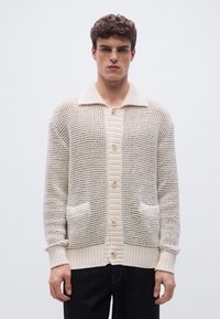 Πλεκτή ζακέτα σε κρεμ απόχρωση με υφή, με γιακά, κουμπώματα και δύο μπροστινές τσέπες πατς.