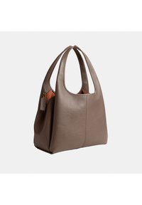 Sac à main en cuir taupe avec une texture douce, des poignées larges et une petite étiquette attachée, présenté sur un fond clair uni.