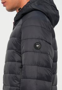 Schwarze Steppjacke mit Kapuze, mit gesteppten Mustern, einem runden Logo-Patch am linken Ärmel und einem teilweise sichtbaren Frontreißverschluss.