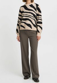 Pull en beige avec des rayures noires façon zèbre, col rond, texture douce et coupe ample, associé à un pantalon large marron foncé et des chaussures noires.