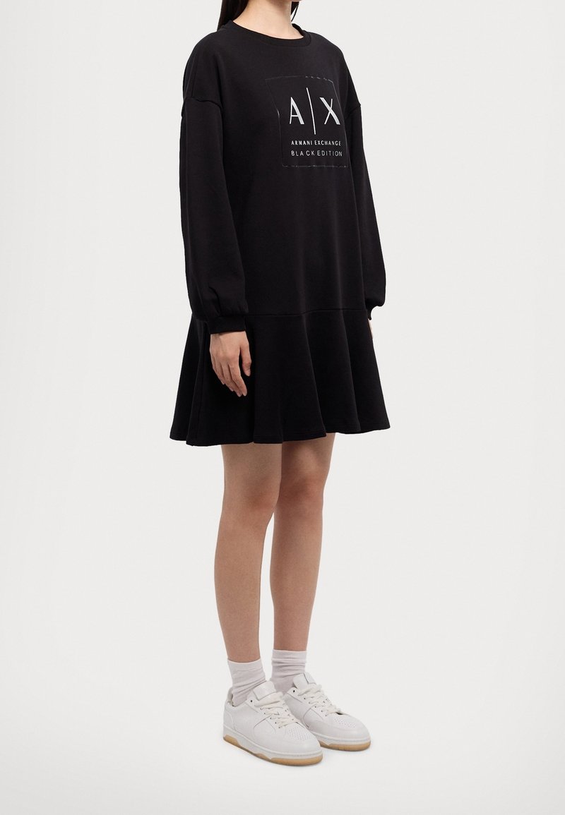 Femme portant une robe sweat Armani Exchange noire à manches longues, avec des chaussettes blanches et des baskets blanches, se tenant sur un fond uni.