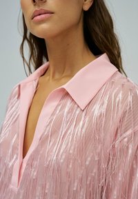 Femme portant un chemisier rose clair avec des détails à franges et un col large, montrant son cou et le bas de son visage sur un fond neutre.