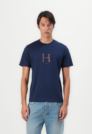 Tamno plava pamučna majica s kratkim rukavima, s velikim burgundskim "H" logotipom u sredini i riječju "Hackett" ispod njega. Klasičan kroj.