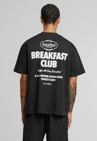 Sort bomulds T-shirt med rund hals, der har fed hvid tekst på ryggen: "Another Breakfast Club" og yderligere detaljer nedenfor.
