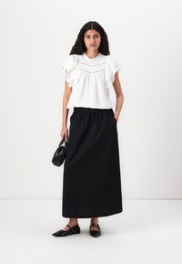 Haut blanc à volants avec des détails texturés, associé à une longue jupe noire avec une taille élastique. Ballerines noires et un petit sac à main noir.