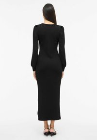 Musta, vartalonmyötäinen neulemekko, jossa on pitkät hihat ja pyöreä pääntie. Mekossa on korostetut olkapäät ja suora, lattiaan ulottuva siluetti.