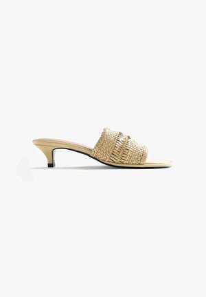 Sandalia mule beige de cuero trenzado con tacón bajo de tipo kitten y puntera abierta, mostrada en perfil lateral sobre fondo blanco.