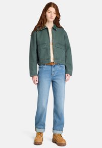 Groene denimjas met kraag en twee voorkruiszakken, gecombineerd met lichtblauwe recht-toe-rechtdoor jeans en tan laarzen.