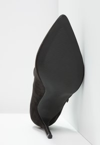 Svart mockaskor med klack, spetig tå och tunn stilettklack. Sulan är texturerad och platt, med en storleksmarkering.