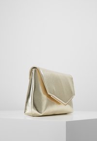 Mała metaliczna złota torebka kopertowa z trójkątnym klapkiem, umieszczona na białej powierzchni na tle jednolitego szarego tła.