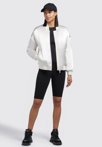 Giacca bomber in satin avorio con polsini e orlo a coste, indossata sopra una maglietta nera e pantaloni corti da bicicletta neri. Un cappellino nero e sneaker nere chunky completano il look.
