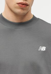 Άνδρας που φοράει γκρι μπλούζα με στρογγυλή λαιμόκοψη και μικρό λευκό λογότυπο New Balance στο αριστερό στήθος, εμφανιζόμενος από τον λαιμ ό έως το πάνω μέρος του στήθους.