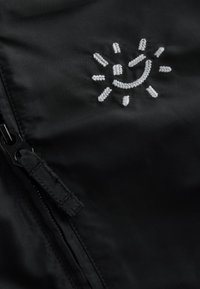 Tissu noir avec un motif de soleil blanc brodé accompagné de rayons. Une fermeture éclair avec une languette de tirage est visible, mettant en valeur le détail du vêtement.