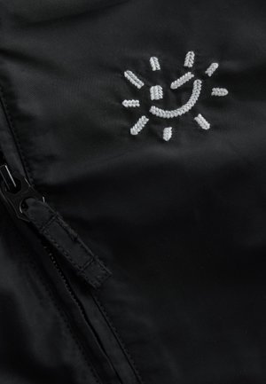 Czarna tkanina z białym haftowanym wzorem słońca z promieniami. Widoczny jest zamek błyskawiczny z uchwytem, podkreślający szczegóły ubrania.