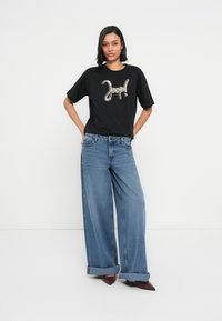 Μαύρο υπερμεγέθες μπλουζάκι με λαμπερό χρυσό σχέδιο που γράφει "Jeep!", σε συνδυασμό με φαρδιά μπλε τζιν παντελόνια και μυτερά μπορντό τακούνια.