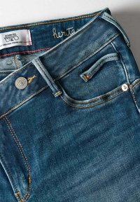 Jean en denim bleu foncé avec une légère décoloration. Les caractéristiques incluent un bouton en métal, des coutures jaunes et une petite poche avant.