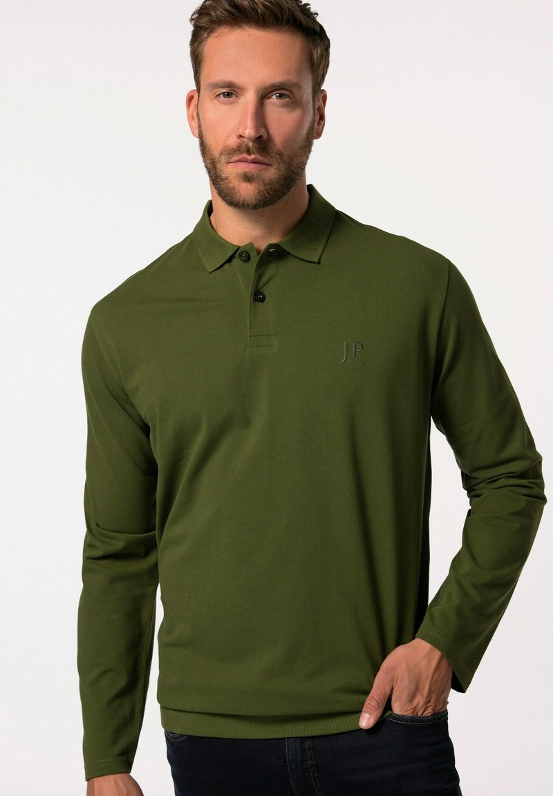 Camisa polo verde de manga larga con un acabado texturizado, tapeta de tres botones y logo bordado en el pecho, combinada con jeans oscuros.