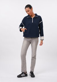 Žena v námorníckom pulóvri s bielymi pruhmi, šedých džínsoch a čiernych teniskách Adidas stojí s jednou rukou v vrecku.