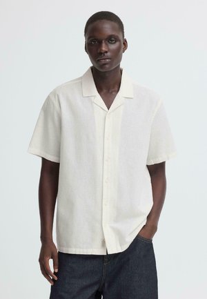 Homme portant une chemise à manches courtes beige clair avec col, boutonnée et non rentrée, sur un jean en denim foncé, posant avec une main dans la poche.