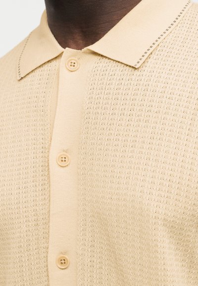 Κοντινό πλάνο ενός ατόμου με σκουρόχρωμη επιδερμίδα που φοράει ένα μπεζ πλεκτό πόλο πουκάμισο με υφή και τρία μπεζ κουμπιά κουμπωμένα.