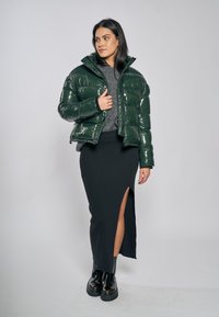 Grön glansig vadderad jacka med hög krage, kombinerad med en lång svart stickad kjol med sidospalt och svarta klumpiga stövlar.