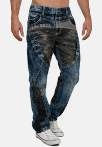 Jean en denim bleu avec des panneaux camouflage, présentant un design taille basse, des coutures décoratives et des accents de boutons sur le côté.