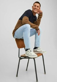 Pull en texture marron et bleu marine, jeans bleu clair et baskets grises. Assis sur une chaise en bois, l'individu sourit avec une main sous le menton.